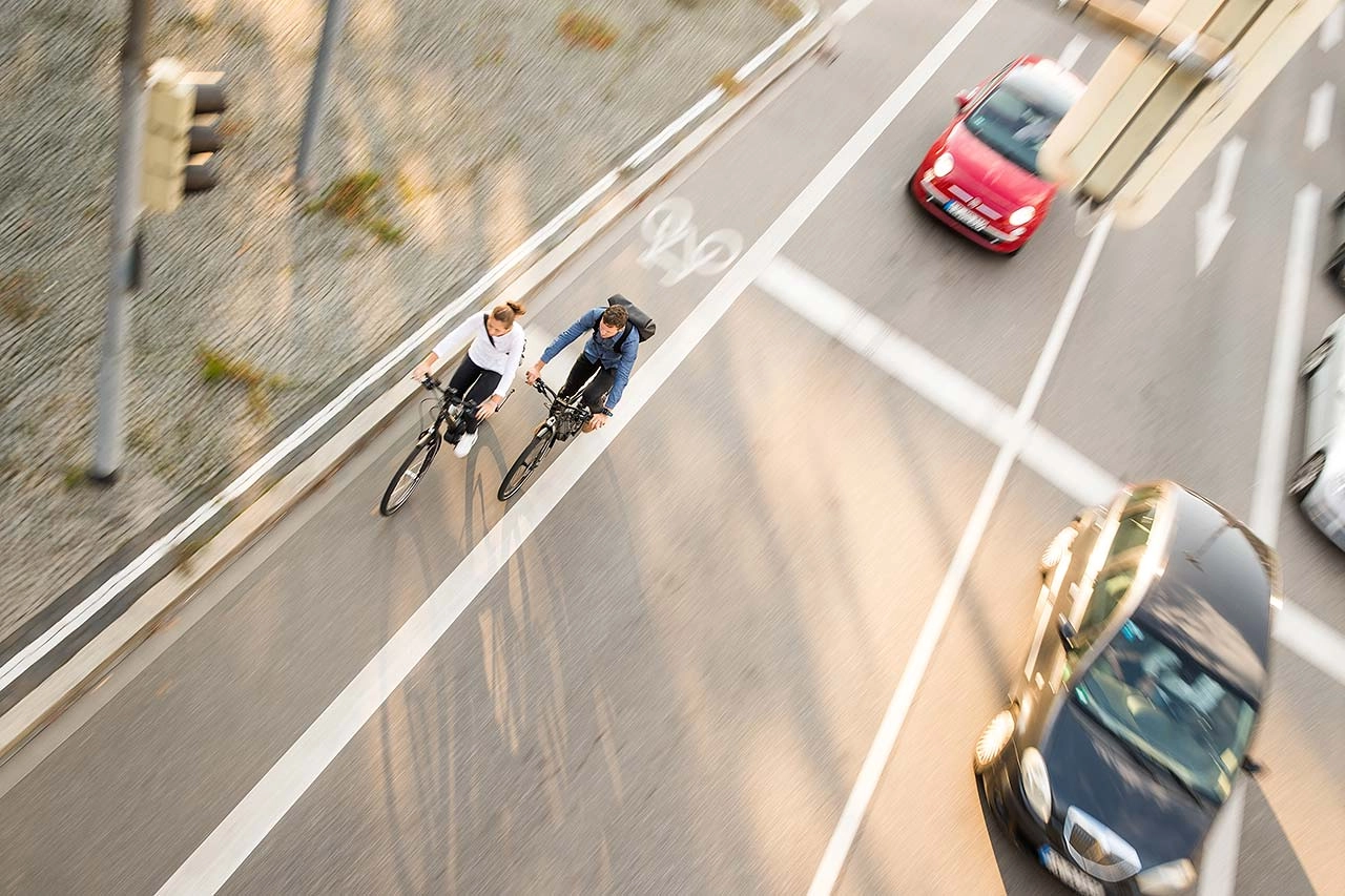 Zwei Fahrradfahrer:innen im Straßenverkehr mit Autos, von schräg vorne/oben fotografiert Zwei Fahrradfahrer:innen im Straßenverkehr mit Autos, von schräg vorne/oben fotografiert