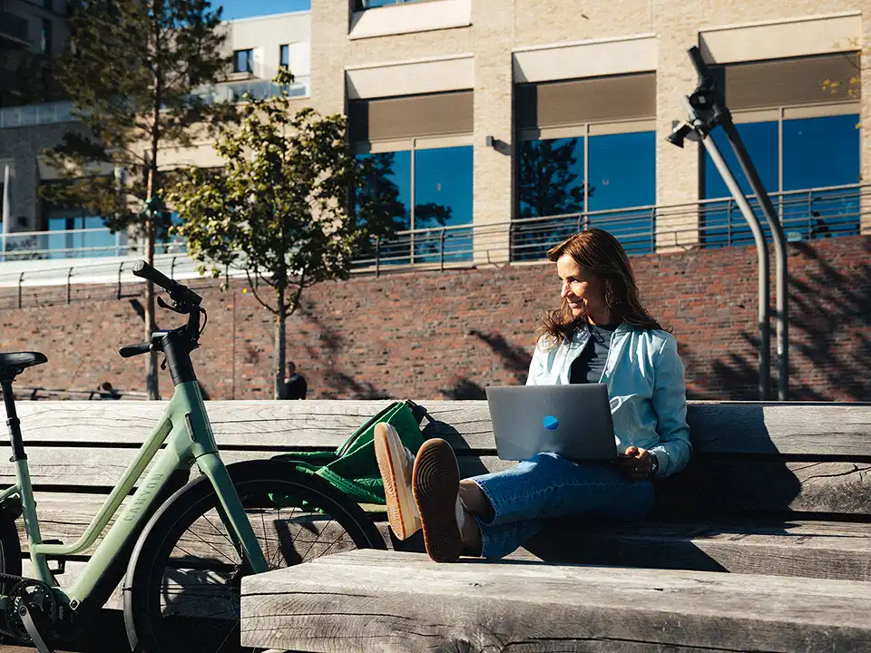 Junge Frau sitzt mit Laptop auf einer Holzbank in der Sonne, neben sich ein E-Bike
